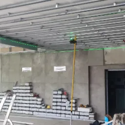 Montaż metalowej konstrukcji sufitu podwieszanego, widoczny laser krzyżowy wyznaczający poziom, ułożone płyty na podłodze, drabiny i narzędzia.