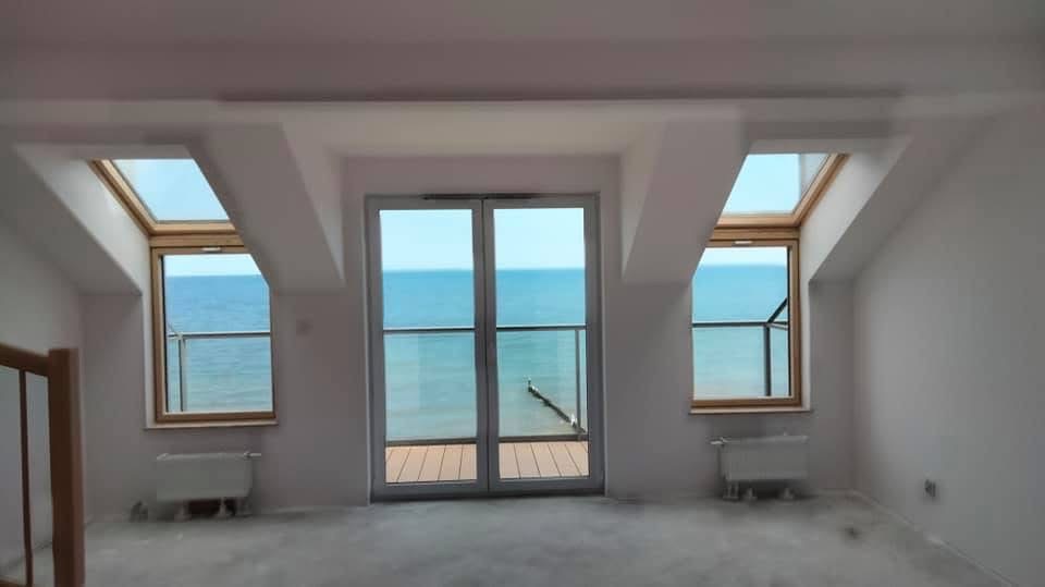 Jasne poddasze z widokiem na morze przez okna dachowe i balkonowe. Świeżo wykończone ściany, grzejniki i surowa podłoga czekają na dalsze prace.