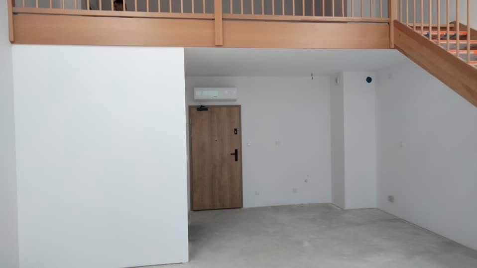 Jasne wnętrze po remoncie: białe ściany, drewniane schody i balustrada antresoli, drzwi wejściowe. Minimalistyczny design, świeżo wykończone pomieszczenie.