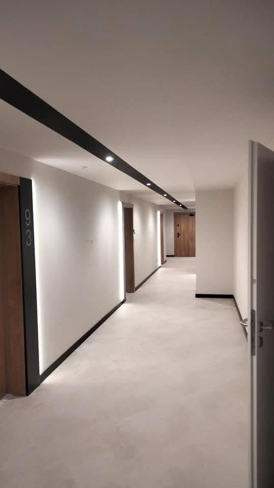 Nowoczesny korytarz w budynku mieszkalnym z białymi ścianami, ciemnymi listwami przypodłogowymi i oświetleniem LED. Widoczne drzwi do mieszkań z numerami.