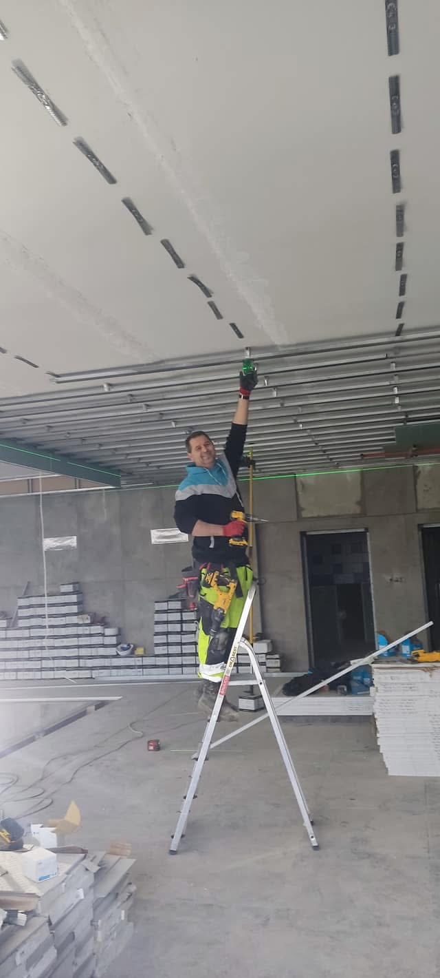 Pracownik na drabinie montuje metalowy stelaż sufitu podwieszanego, używając poziomicy laserowej. W tle stosy płyt gipsowo-kartonowych w surowym wnętrzu.