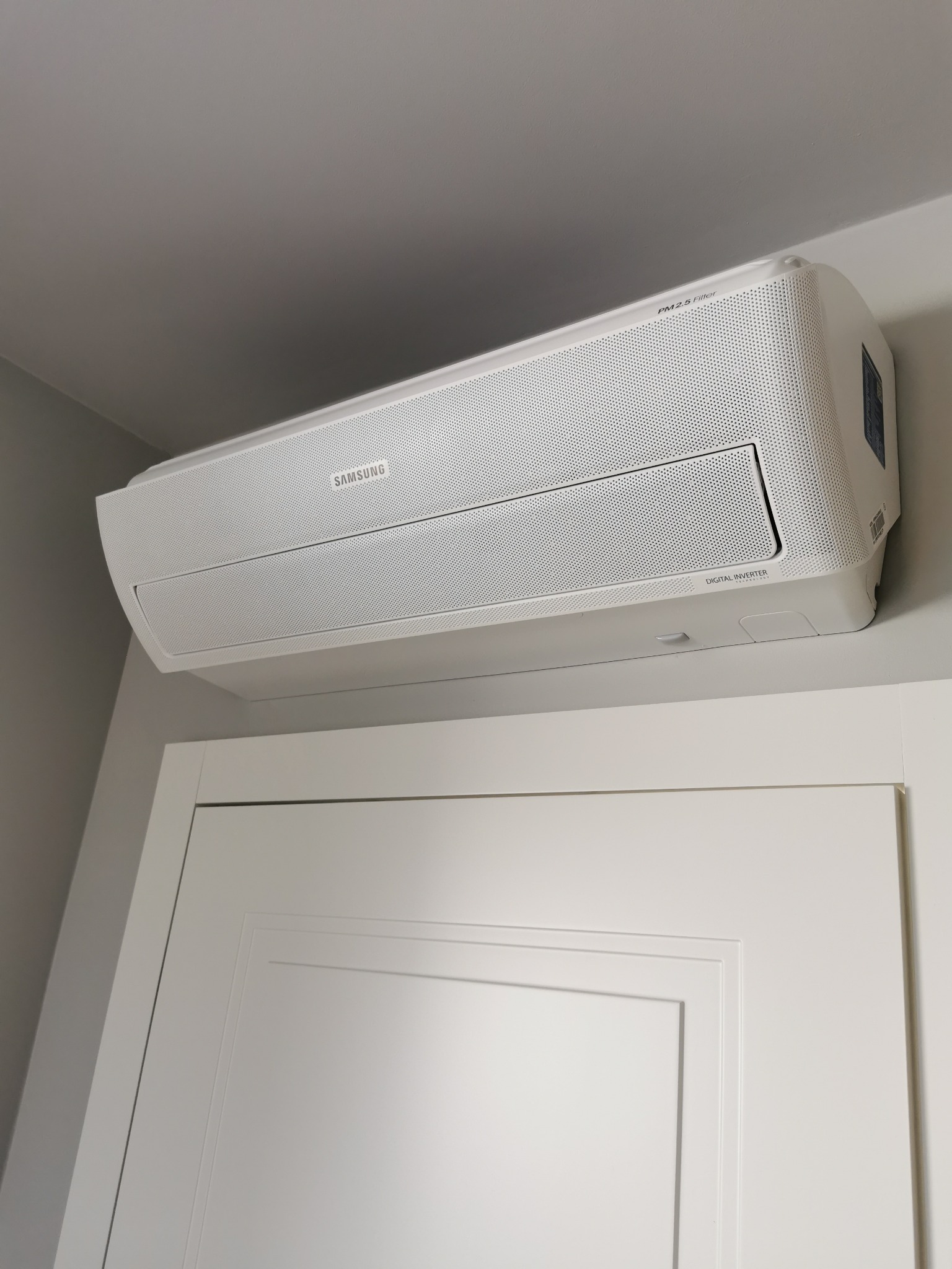 Biała jednostka klimatyzacji Samsung Digital Inverter zamontowana nad białą, panelową wewnętrzną stroną drzwi w pomieszczeniu o szarych ścianach.