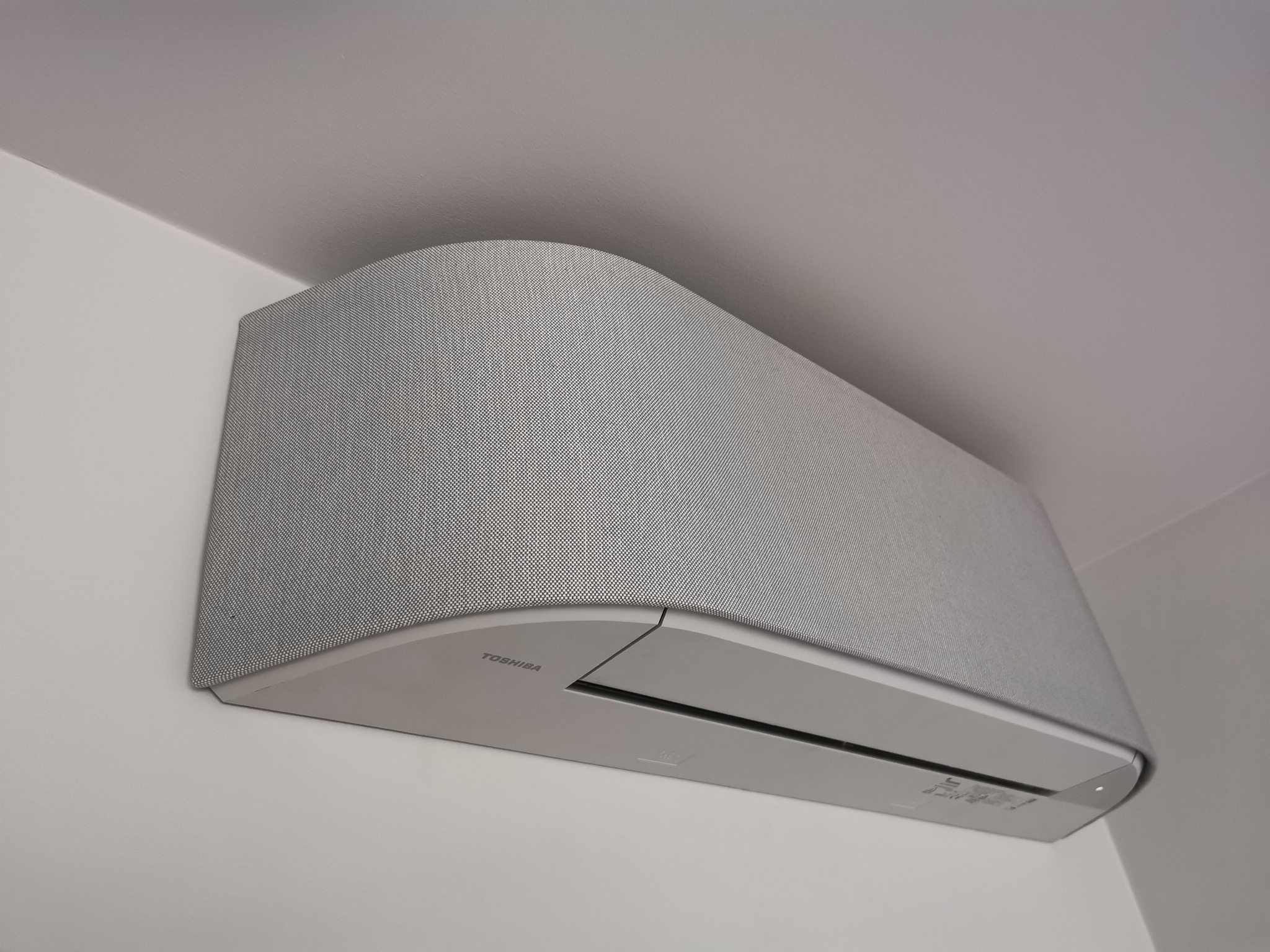 Szara klimatyzacja Toshiba zamontowana na ścianie, z obudową imitującą tkaninę, w minimalistycznym wnętrzu.