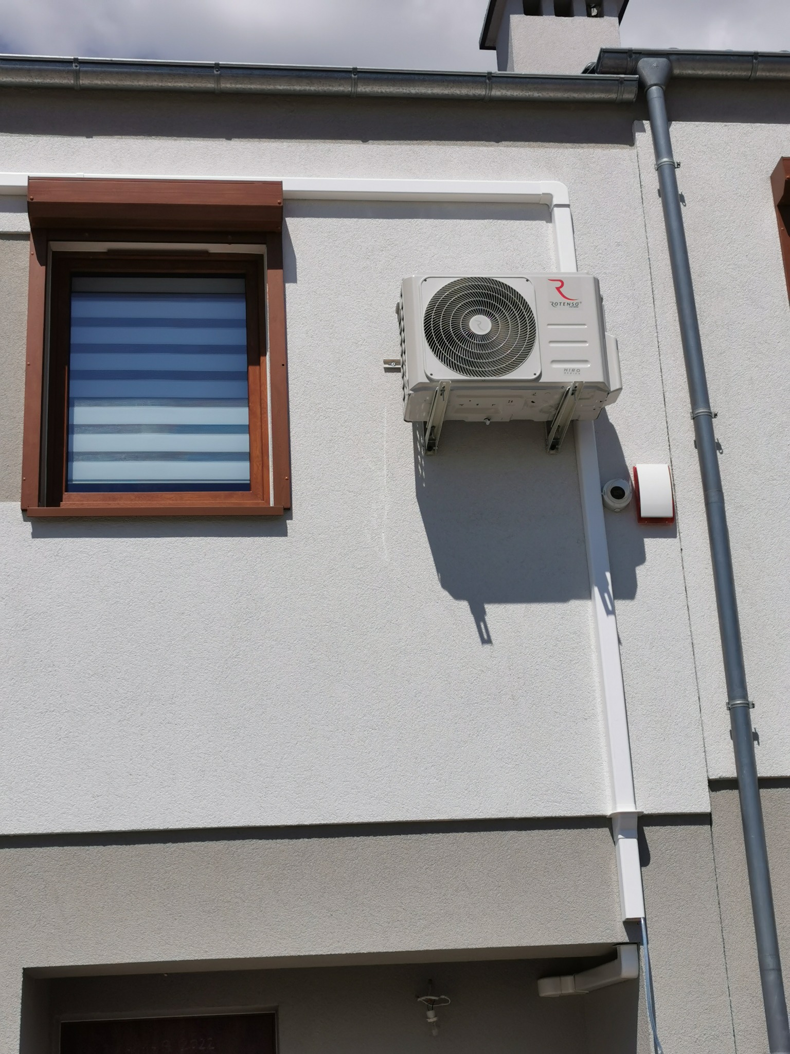 Zewnętrzna jednostka klimatyzacji Rotenso Hiro zamontowana na elewacji domu, widoczne białe korytka instalacyjne, okno z brązową ramą i żaluzjami oraz rynna.