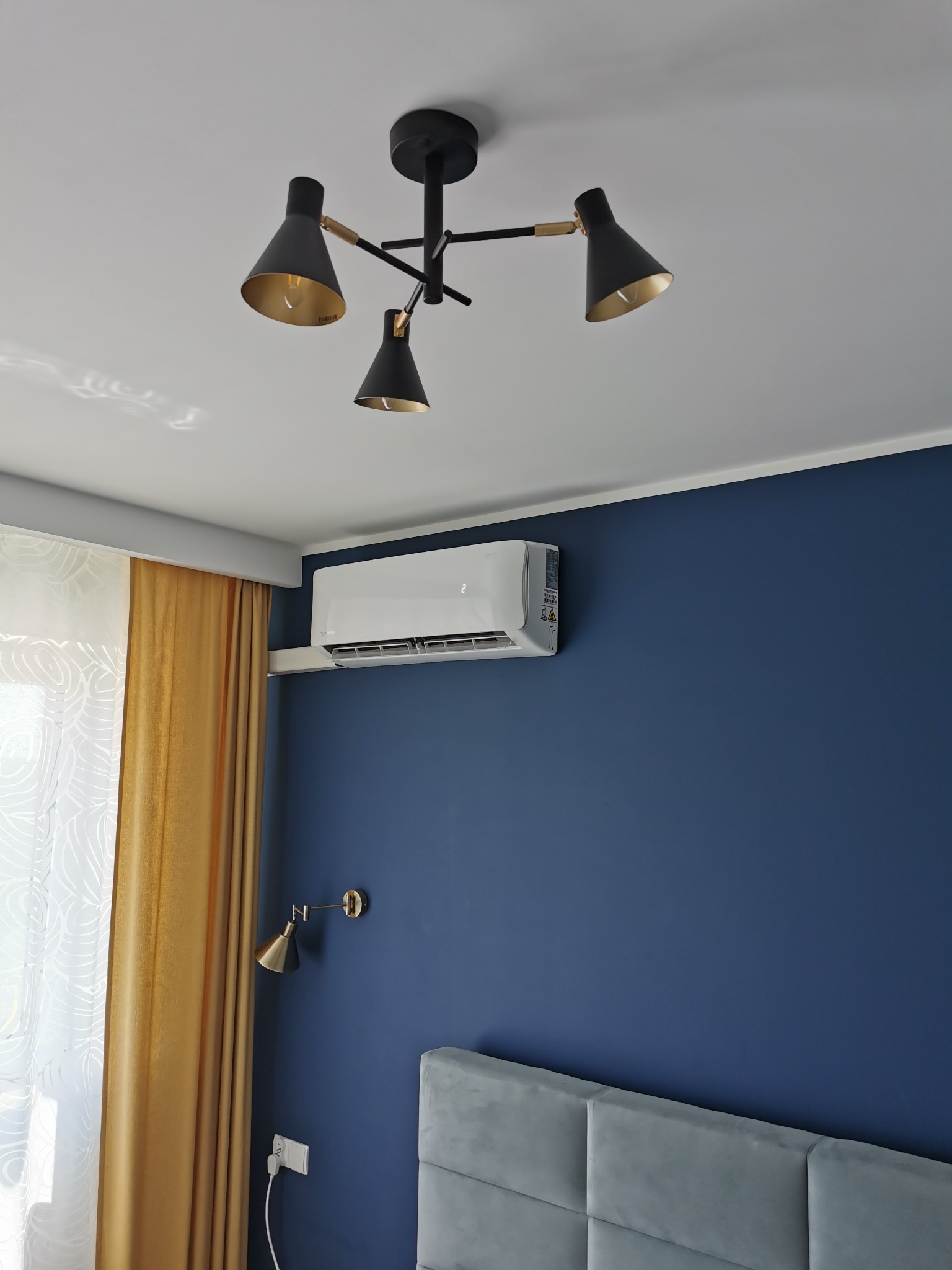 Nowoczesny klimatyzator ścienny zamontowany w sypialni w Poznaniu, widoczny na tle granatowej ściany, pod czarną lampą sufitową z elementami złotymi.