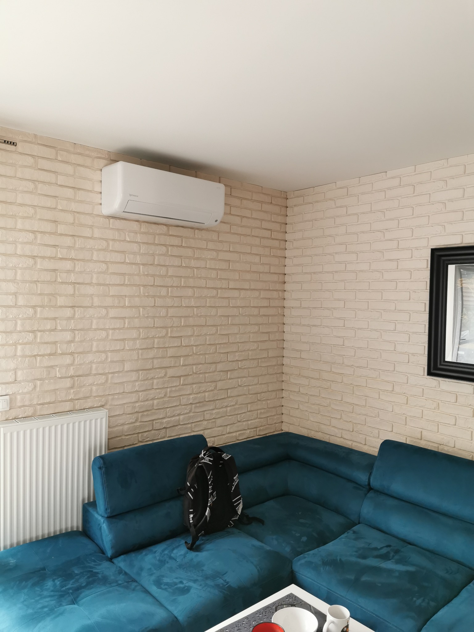 Biały klimatyzator ścienny zamontowany w rogu pokoju z ceglaną ścianą, widoczny fragment niebieskiej sofy i plecak leżący na niej.
