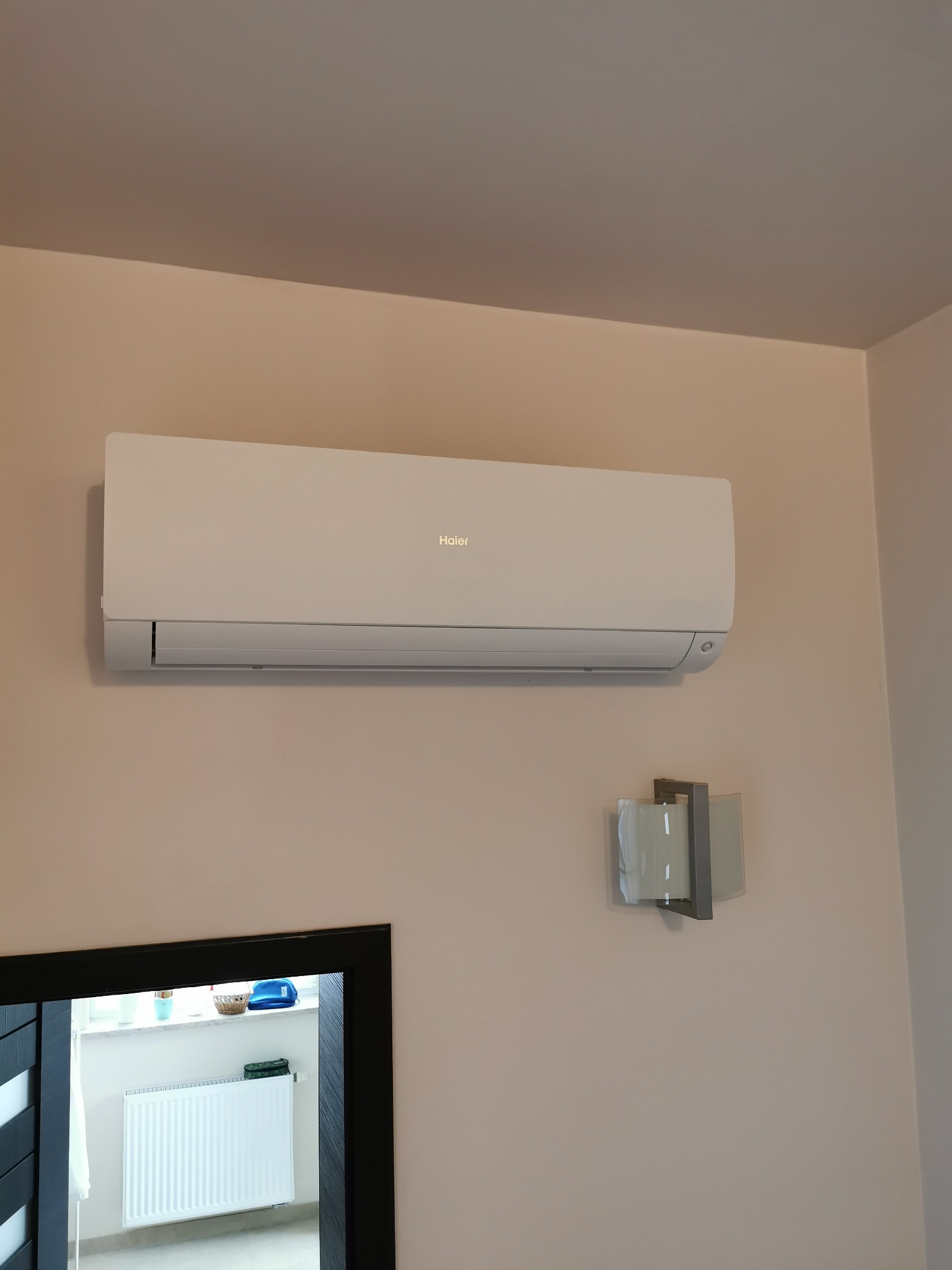 Klimatyzator Haier zamontowany na ścianie w pokoju, widoczny fragment lustra z odbiciem grzejnika i okna, obok lampka ścienna.