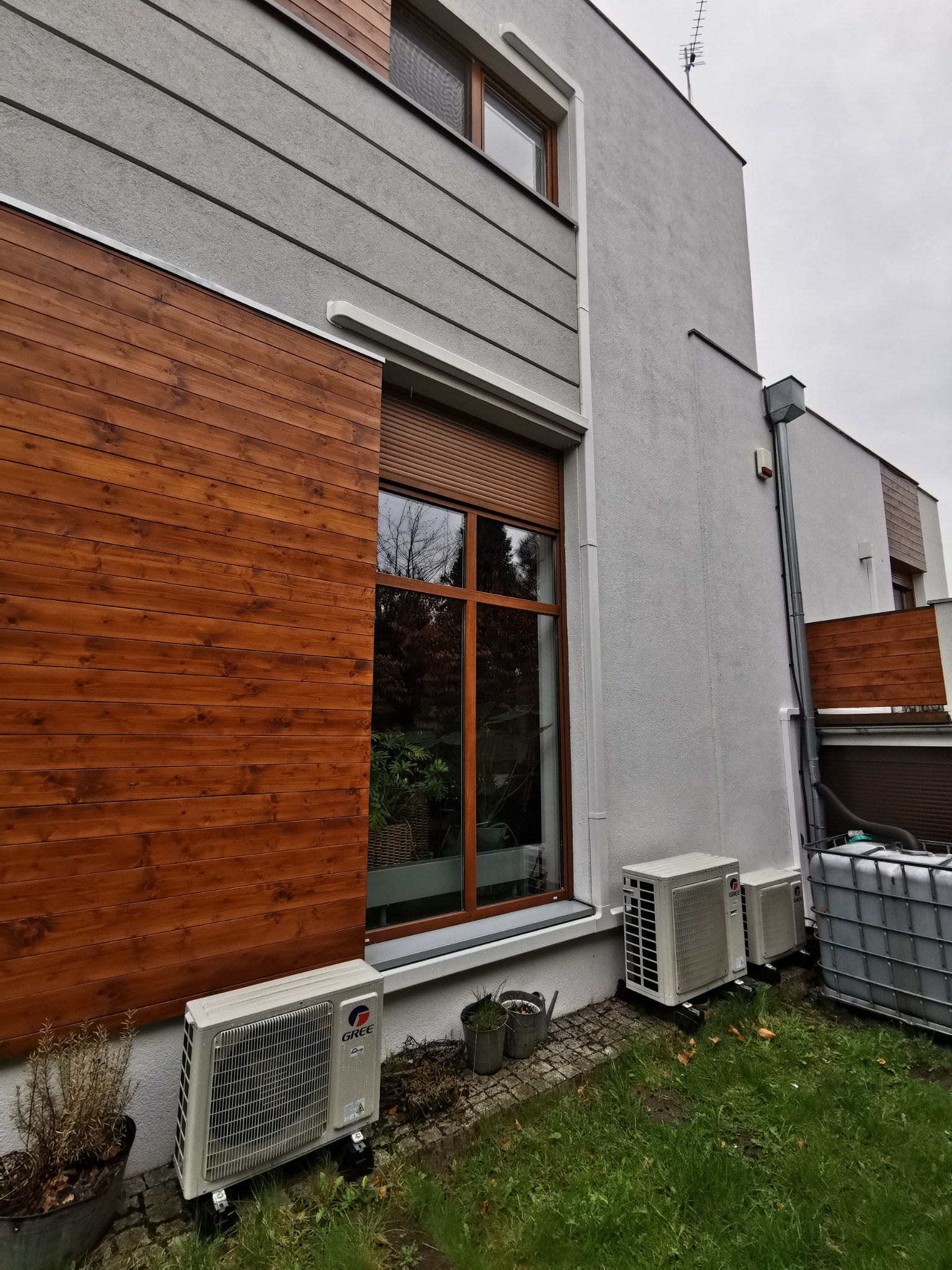 Zewnętrzne jednostki klimatyzacji marki Gree zamontowane przy nowoczesnym domu z drewnianą elewacją i dużym oknem z brązowymi ramami, widoczne elementy instalacji odprowadzającej wodę.