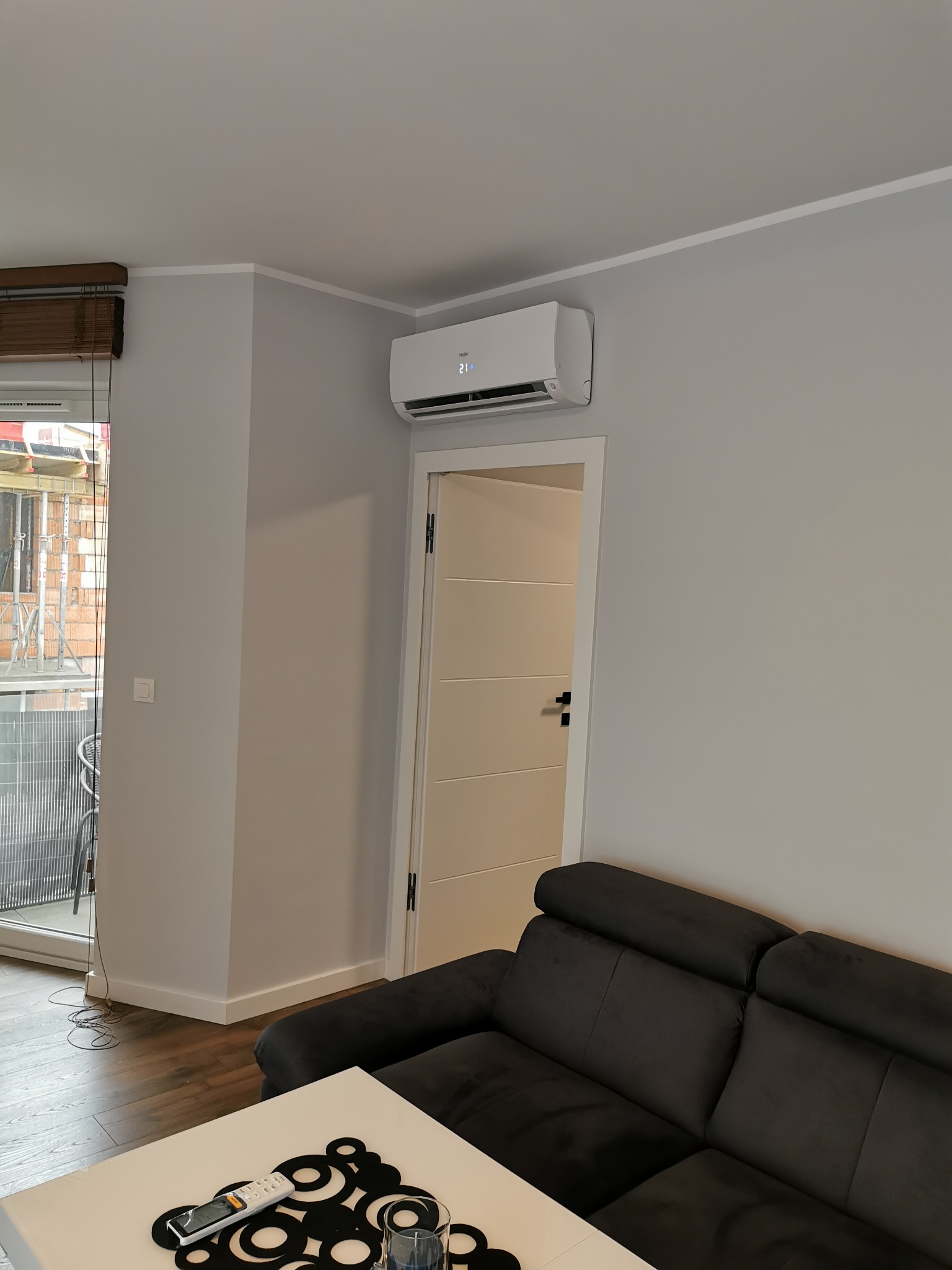 Biały klimatyzator ścienny w szarym salonie z balkonem, widoczny pilot i dekoracje na stole.