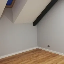 Poddasze po remoncie: drewnopodobne panele podłogowe, szara ściana z białą listwą przypodłogową, okno dachowe z widokiem na błękitne niebo, czarna belka stropowa.