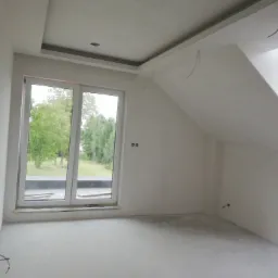 Poddasze po szpachlowaniu i gładziowaniu, z widocznymi przewodami elektrycznymi wystającymi ze ściany, przygotowane do dalszych prac wykończeniowych. Okno z widokiem na zieleń.