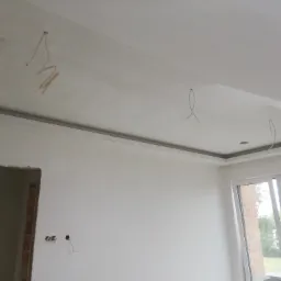 Wnętrze w trakcie remontu: sufit podwieszany z płyty karton gips z widocznymi przewodami elektrycznymi, ściany przygotowane do malowania, okno z widokiem na zieleń.