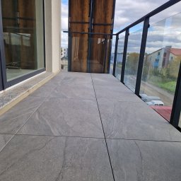 PERFECT HOUSE Wykończenia Wnętrz Adam Szypot - Szare, matowe płytki gresowe ułożone na balkonie z przeszkloną balustradą. Widok na zabudowę osiedlową i błękitne niebo z chmurami w tle.