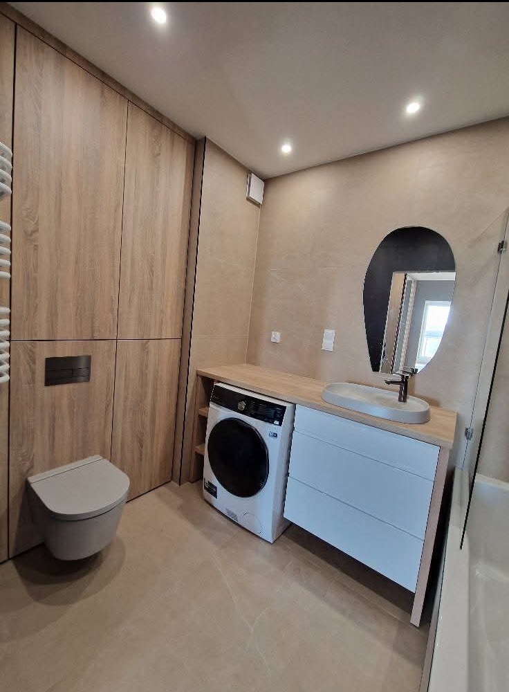 Nowoczesna łazienka z zabudową meblową w odcieniach drewna, pralką pod blatem i asymetrycznym lustrem. Uwagę zwraca minimalistyczna toaleta podwieszana.