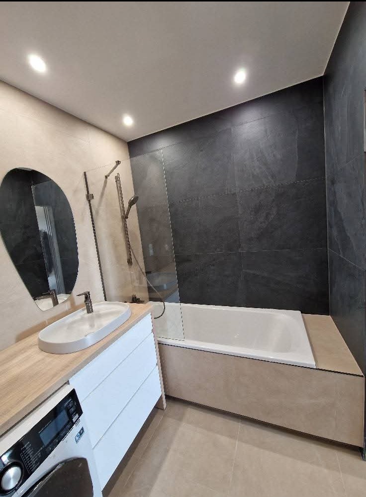 Nowoczesna łazienka z owalnym lustrem, umywalką nablatową i kabiną prysznicową typu walk-in. Ciemne płytki na ścianie kontrastują z jasną podłogą i drewnianym blatem.