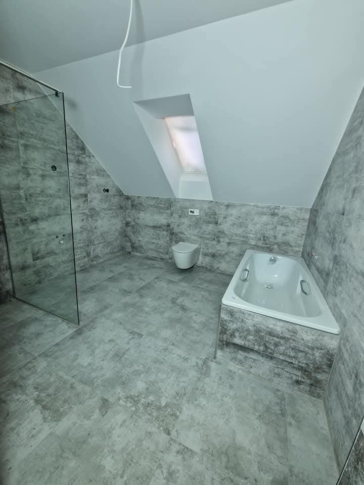 Szara łazienka na poddaszu ze szklaną kabiną, toaletą i wanną w zabudowie. Płytki imitujące beton na ścianach i podłodze. Okno dachowe w skosie.