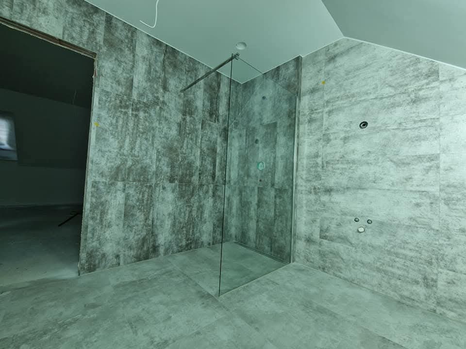 Nowoczesna kabina prysznicowa ze szklaną ścianką i szarymi płytkami imitującymi beton, widoczne przygotowane punkty instalacji wodnej. Minimalistyczny design.