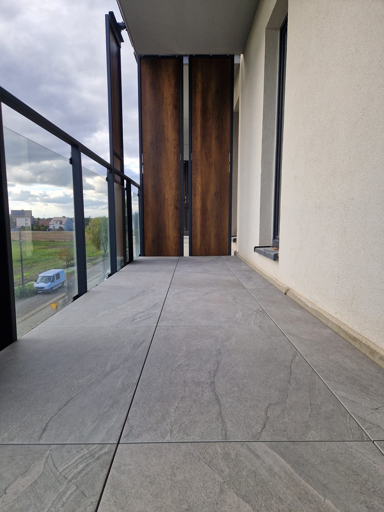 Nowoczesny balkon z szarymi płytkami, szklaną balustradą i drewnianymi panelami. Widok na okolicę z błękitnym busem w tle. Minimalistyczny design.