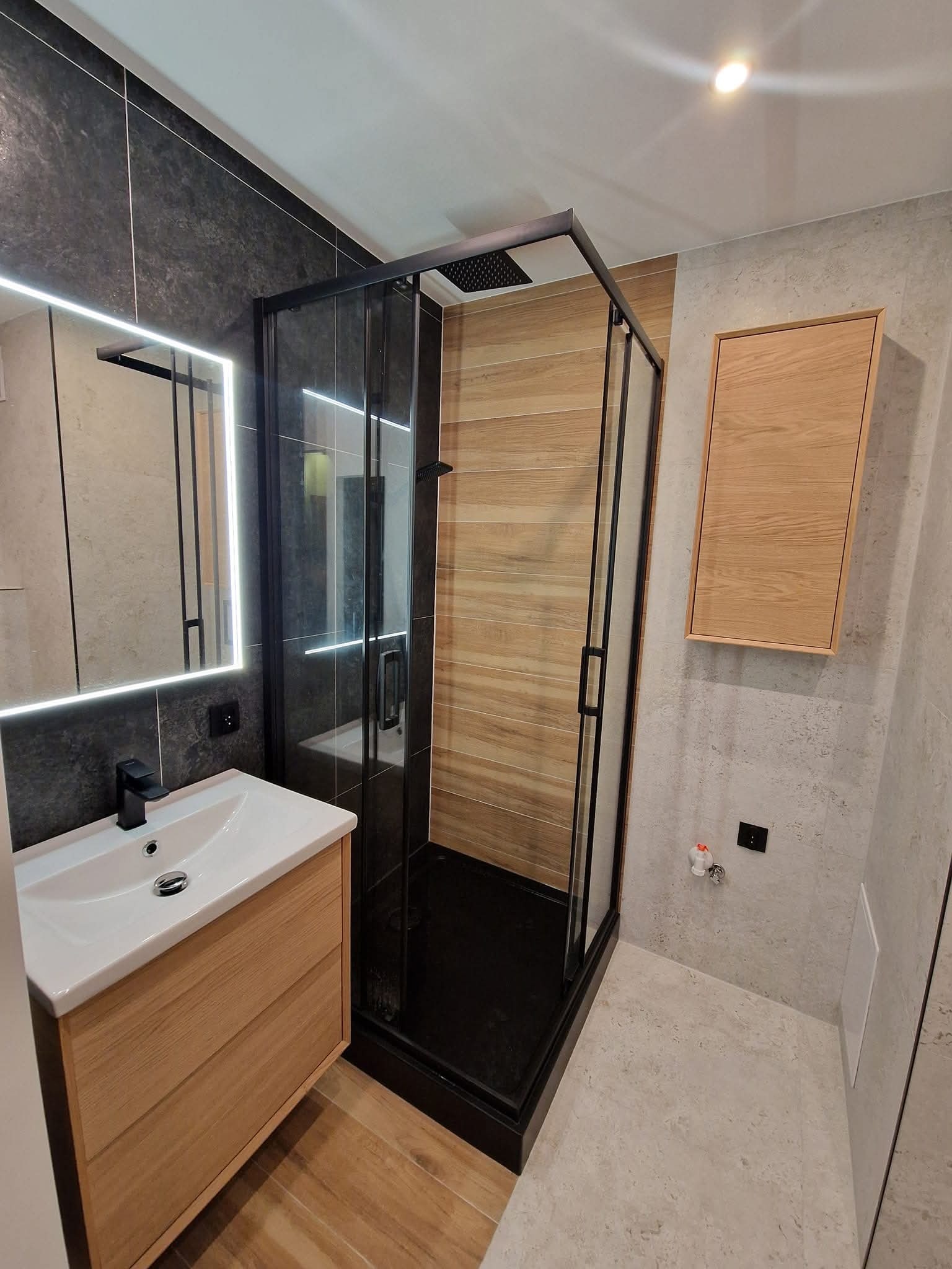 Nowoczesna łazienka z kabiną prysznicową typu walk-in z czarnymi ramami, drewnianą szafką pod umywalką i podświetlanym lustrem. Ściany wykończone płytkami w dwóch odcieniach szarości.