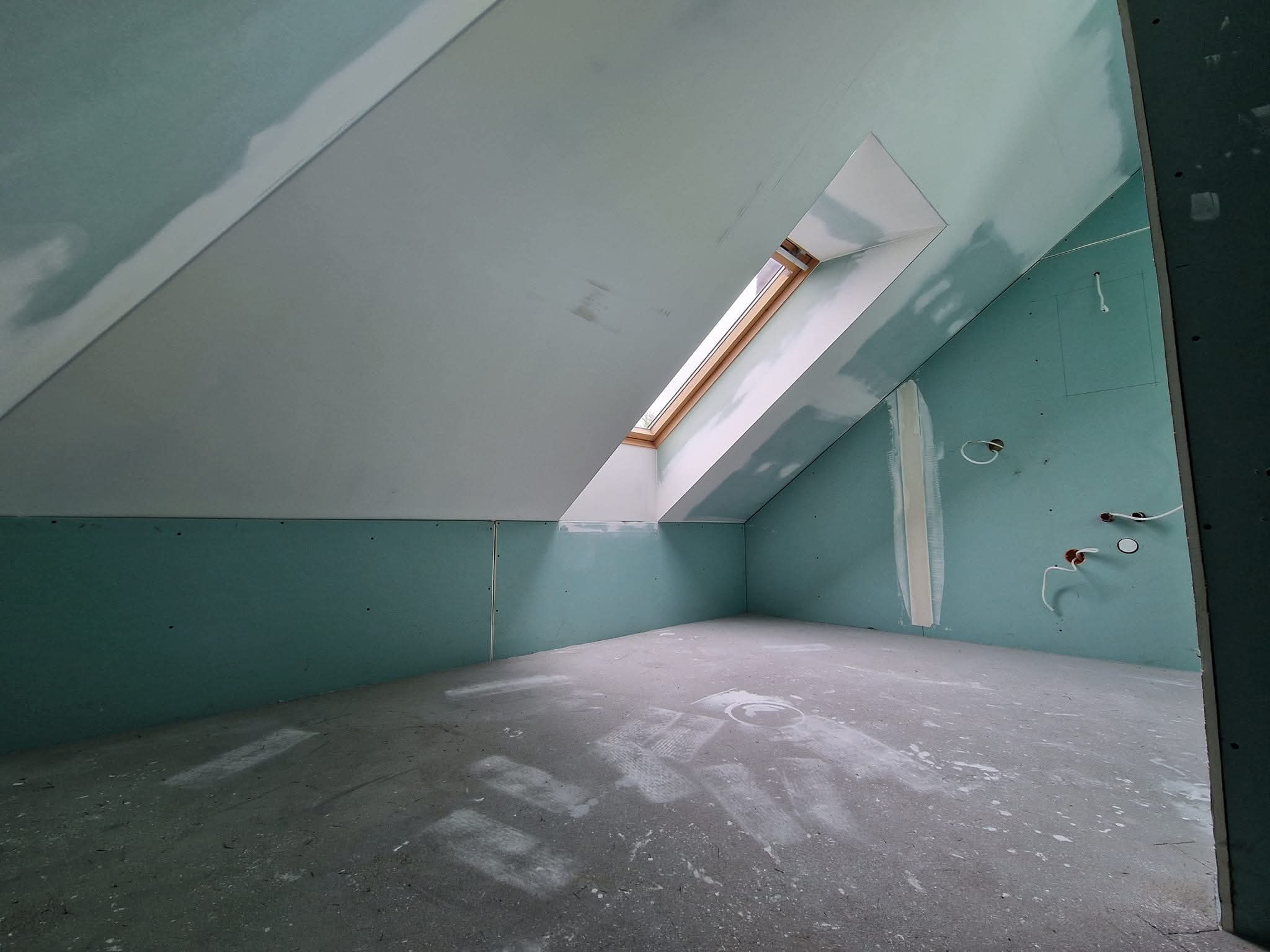 Poddasze w trakcie wykończenia: płyty gipsowo-kartonowe na ścianach i skosach, okno dachowe, widoczne instalacje elektryczne. Ślady gładzi na podłodze.