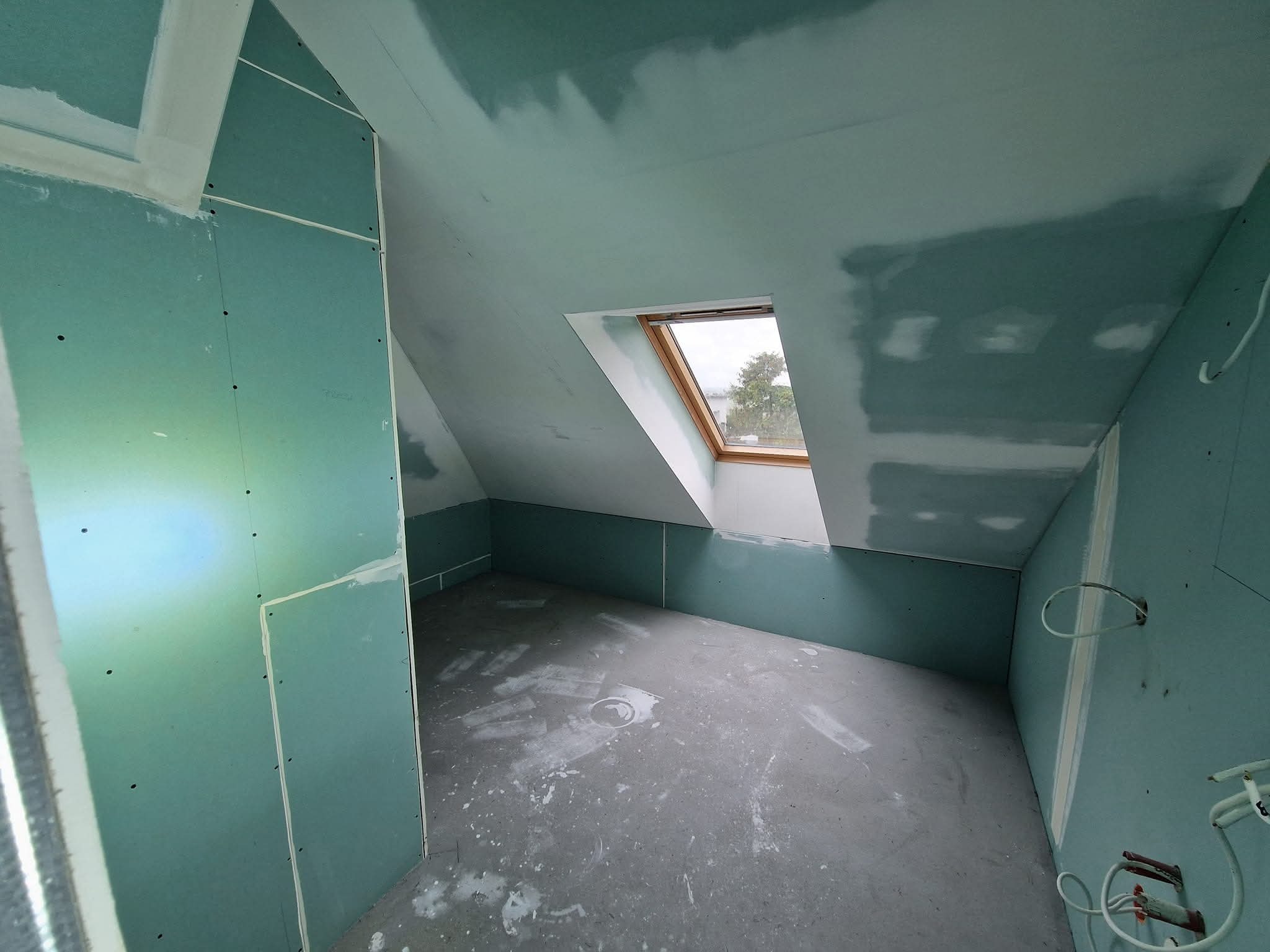 Poddasze w trakcie adaptacji: ściany i skosy obłożone płytami gipsowo-kartonowymi w kolorze zielonym, widoczne łączenia i szpachlowanie, okno dachowe. Surowy stan.