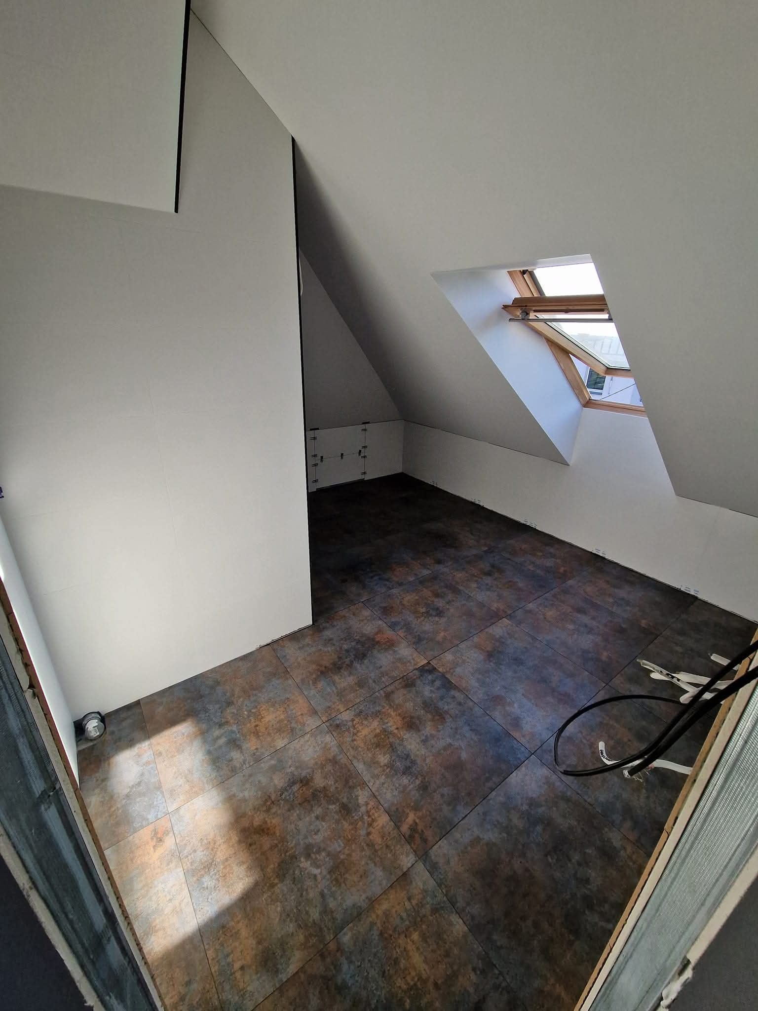 Poddasze po remoncie: brązowo-szara płytki podłogowe, białe ściany, okno dachowe. Widoczne narzędzia i kable. Ukończone wykończenie wnętrza.