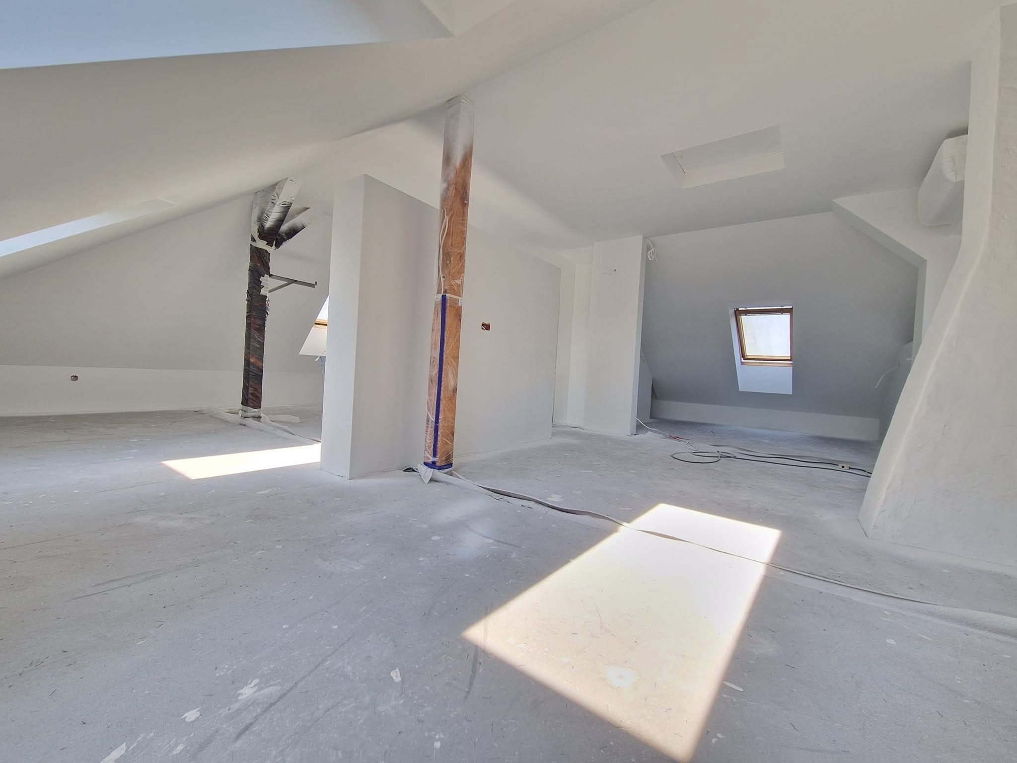 Jasne poddasze po malowaniu na biało, z widocznymi promieniami słońca wpadającymi przez okna dachowe i zabezpieczonymi folią kolumnami. Świeżo pomalowane ściany i podłoga.