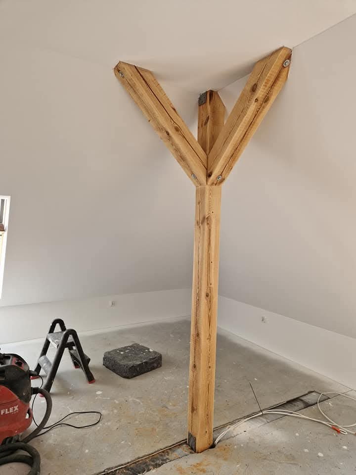 Drewniana konstrukcja nośna w kształcie litery Y, wspierająca skośny sufit poddasza. Widoczny odkurzacz przemysłowy i drabina, sugerujące prace wykończeniowe.