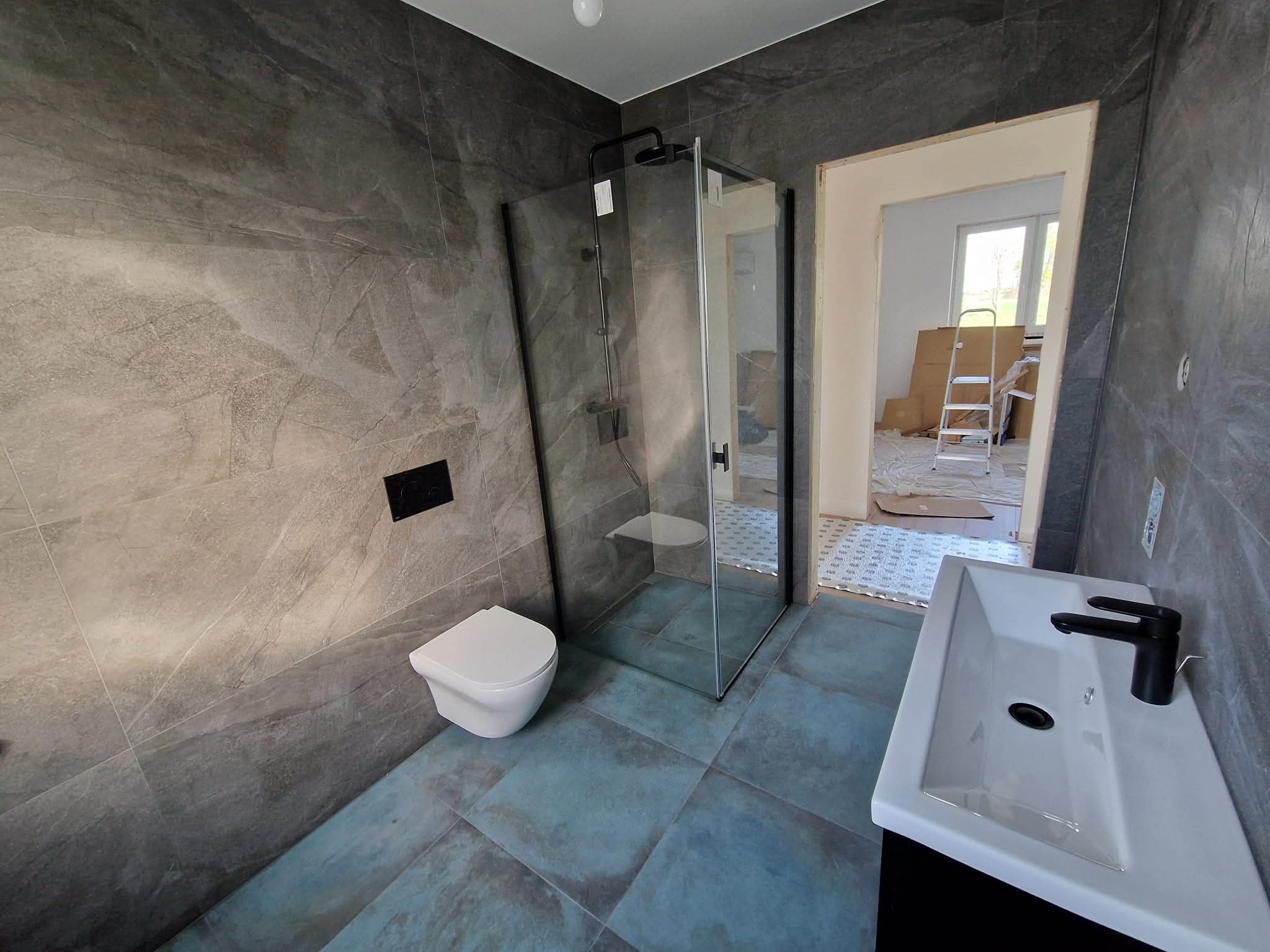 Wykończona łazienka z szarymi płytkami imitującymi kamień na ścianach, kabiną prysznicową typu walk-in i umywalką z czarną armaturą. Na podłodze niebieskie płytki.