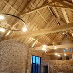 Wnętrze stodoły z odsłoniętą drewnianą konstrukcją dachu, oświetlone nowoczesnymi okrągłymi lampami wiszącymi, z ceglanymi ścianami i dużym oknem w tle.