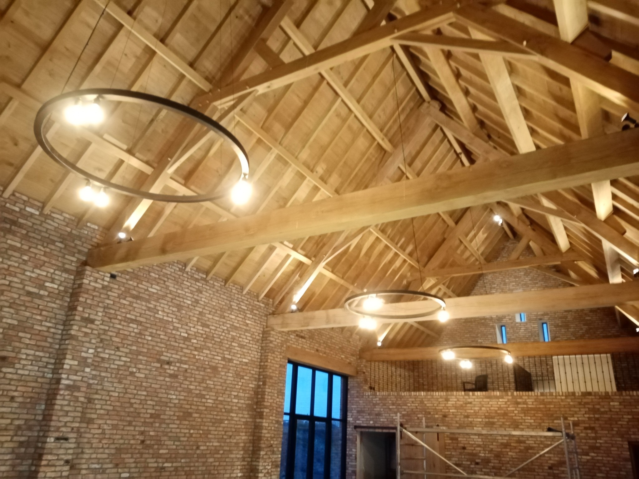 Wnętrze stodoły z odsłoniętą drewnianą konstrukcją dachu, oświetlone nowoczesnymi okrągłymi lampami wiszącymi, z ceglanymi ścianami i dużym oknem w tle.