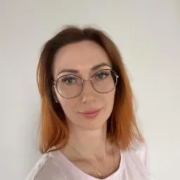 Portret kobiety w okularach, z rudymi włosami, uśmiechniętej, na jasnym tle.