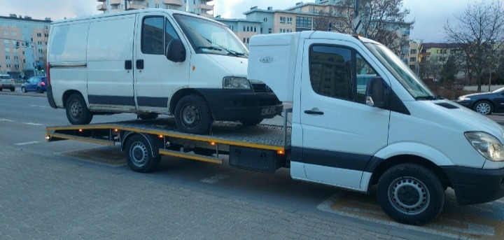 Biała laweta przewozi białego dostawczaka na tle miejskiej ulicy w pochmurny dzień.