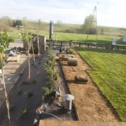 Fragment ogrodu w trakcie zakładania: sadzonki drzewek, rozkładana trawa z rolki, system nawadniania, czarna agrowłóknina, widok z góry na posesję z domami i polem w tle.