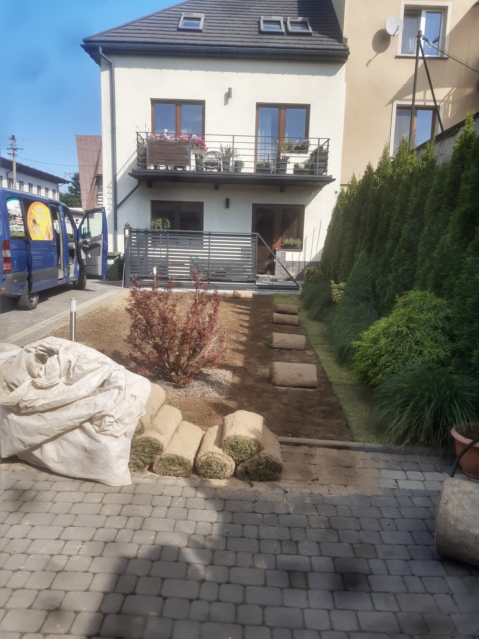Rozwinięte rolki trawy w rzędzie na przygotowanym podłożu, widoczny fragment trawnika, budynek mieszkalny z balkonami w tle, niebieski samochód dostawczy z otwartymi drzwiami po lewej stronie.