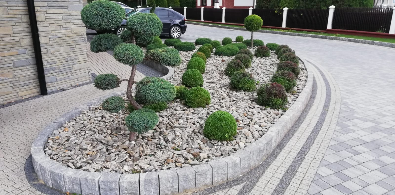 Ogród z ozdobnymi krzewami w formie kul, wysypany kamieniami, obrzeże z kostki, w tle podjazd z kostki i samochód.