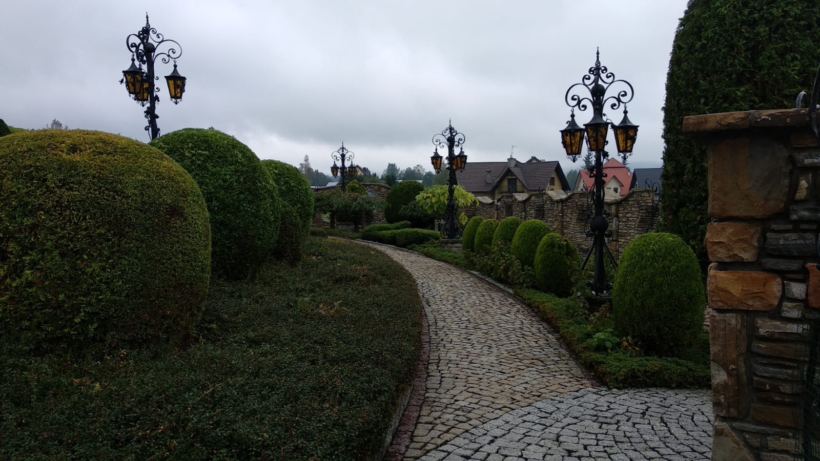 Urokliwa ścieżka z kostki brukowej w ogrodzie z równo przyciętymi krzewami i ozdobnymi latarniami, prowadząca w kierunku zabudowań w tle.