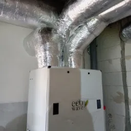 Biała jednostka rekuperatora Eco Wave z podłączonymi, zaizolowanymi przewodami wentylacyjnymi w pomieszczeniu z surowymi ścianami z betonu komórkowego.