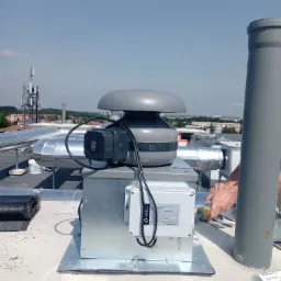 Montaż wentylatora dachowego z szarym grzybkiem na dachu budynku komercyjnego, widoczne ręce uszczelniające silikonem, w tle zabudowa miejska i maszt telekomunikacyjny.