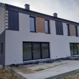 Dwukondygnacyjny dom w trakcie prac elewacyjnych, z fragmentami ścian w kolorze szarym i białym, widoczne drewniane elementy konstrukcyjne i czarne panele na elewacji, teren wokół domu w trakcie...