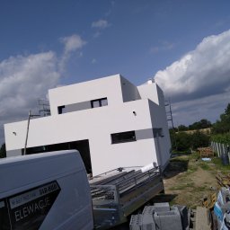 JAR BUD - Nowoczesny dom z białą elewacją i szarymi akcentami, widoczny fragment firmowego samochodu z napisem 'Elewacje' oraz przyczepa z materiałami budowlanymi, na tle błękitnego nieba z chmurami.