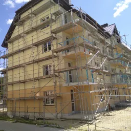 Wielopiętrowy budynek w trakcie ocieplania elewacji, widoczne rusztowania i dwukolorowa elewacja - górna część jasna, dolna w odcieniach beżu.