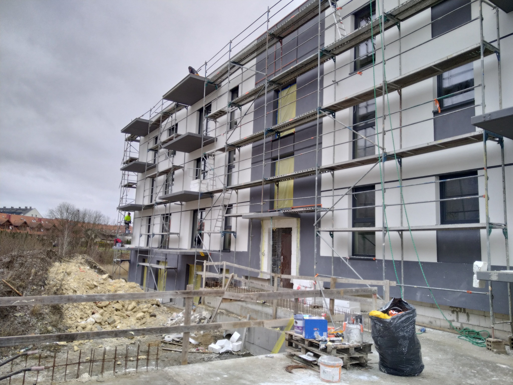 Wielopiętrowy budynek w trakcie prac elewacyjnych, widoczne rusztowania, fragmenty ocieplenia i pracownicy na różnych poziomach, teren budowy z gruzem i narzędziami.
