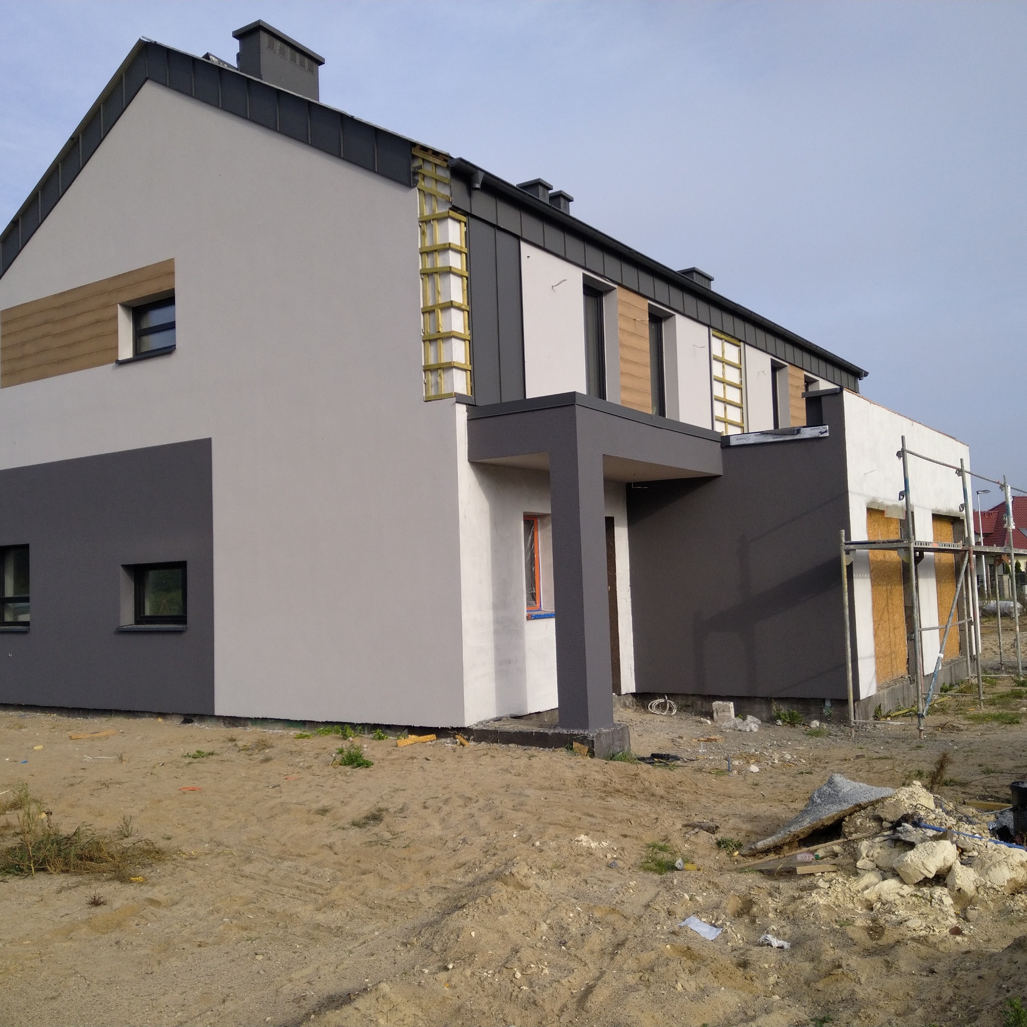 Dwukondygnacyjny dom w trakcie prac elewacyjnych, widoczne fragmenty ocieplenia i rusztowania, częściowo pomalowany na szaro i biało, z elementami drewnianymi.