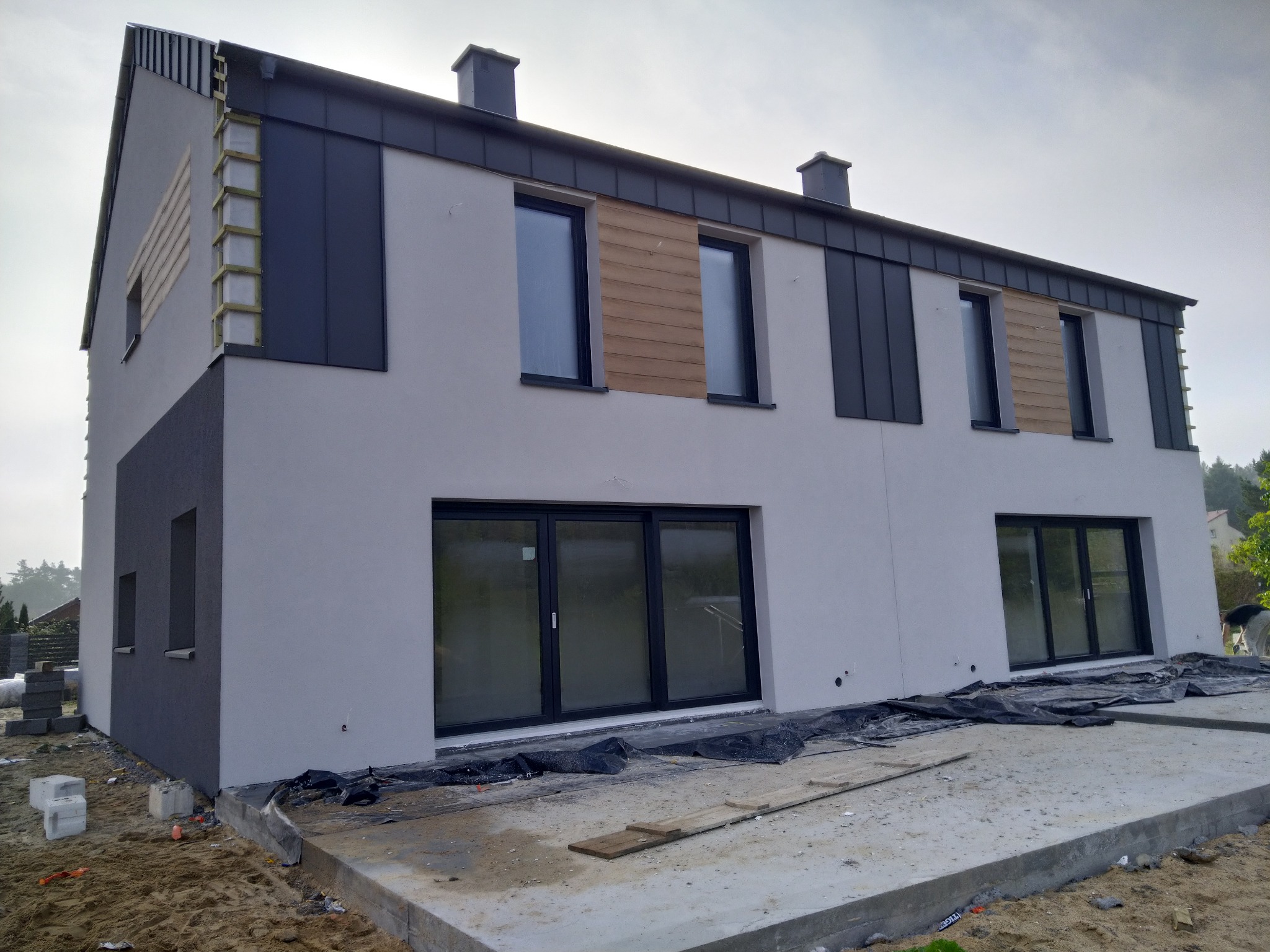 Dwukondygnacyjny dom w trakcie prac elewacyjnych, z fragmentami ścian w kolorze szarym i białym, widoczne drewniane elementy konstrukcyjne i czarne panele na elewacji, teren wokół domu w trakcie...