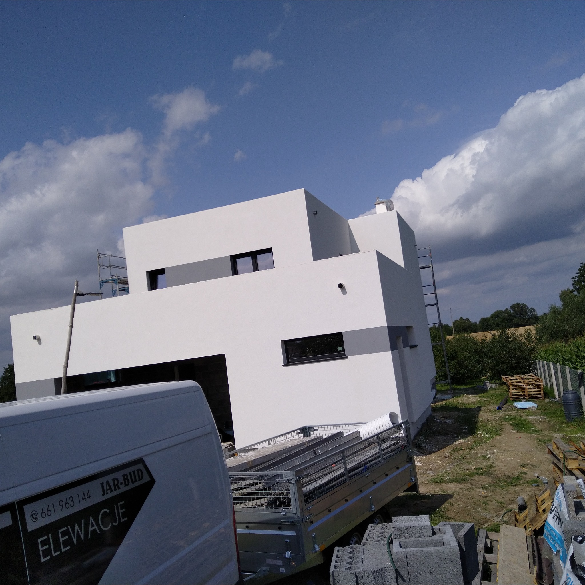 Nowoczesny dom z białą elewacją i szarymi akcentami, widoczny fragment firmowego samochodu z napisem 'Elewacje' oraz przyczepa z materiałami budowlanymi, na tle błękitnego nieba z chmurami.