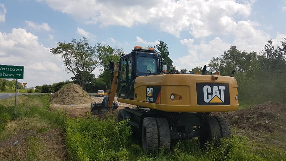 Żółta koparka CAT M318D na poboczu drogi z drogowskazem na Warszawę i Puławy, obok pryzma ziemi i wykop