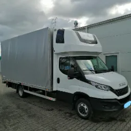 Biały samoch&oacute;d dostawczy Iveco Daily z plandeką na parkingu z kostki brukowej w pochmurny dzień.