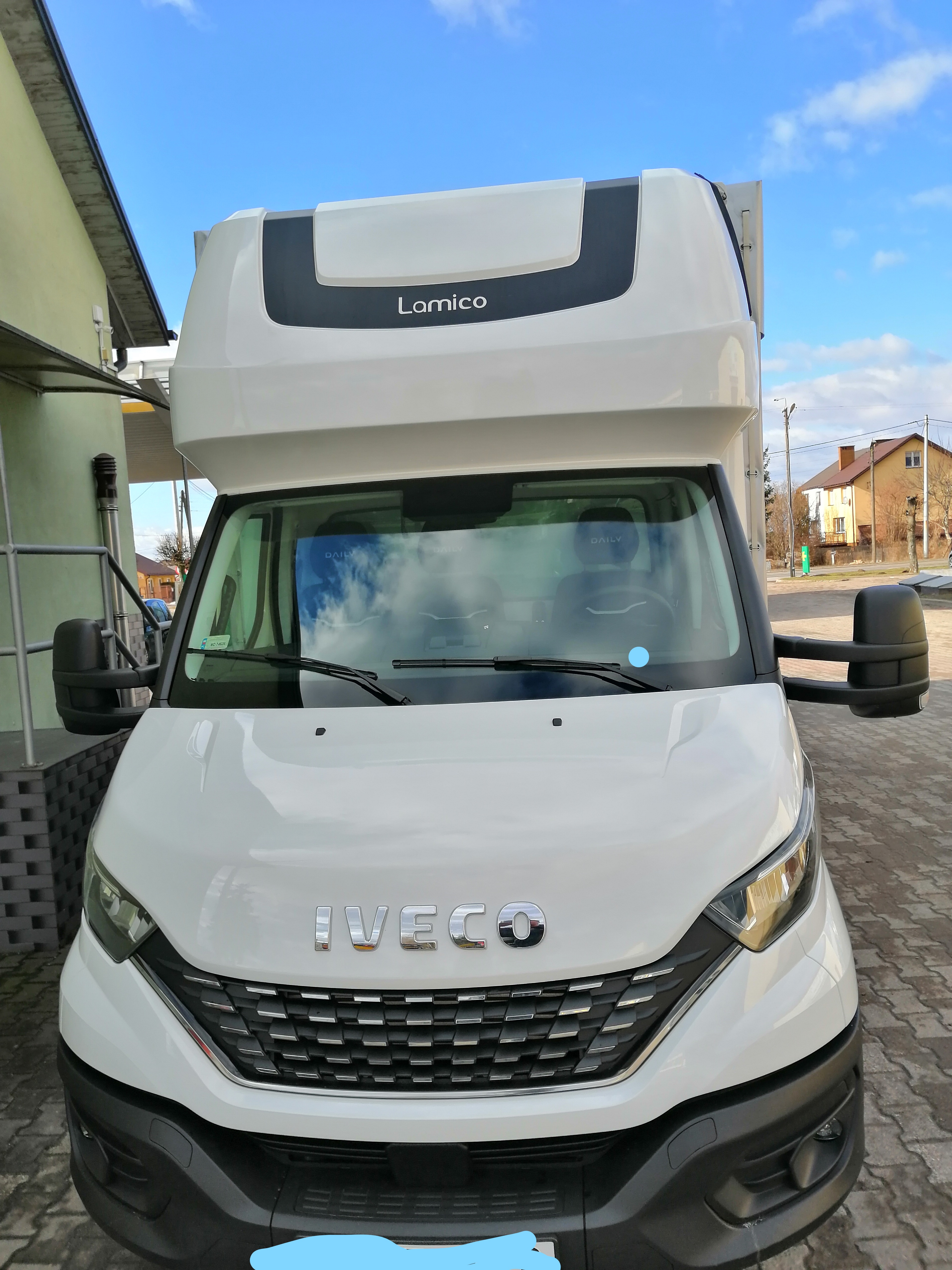 Biały bus Iveco Lamico z wysoką kabiną, widok z przodu na tle budynku i nieba.