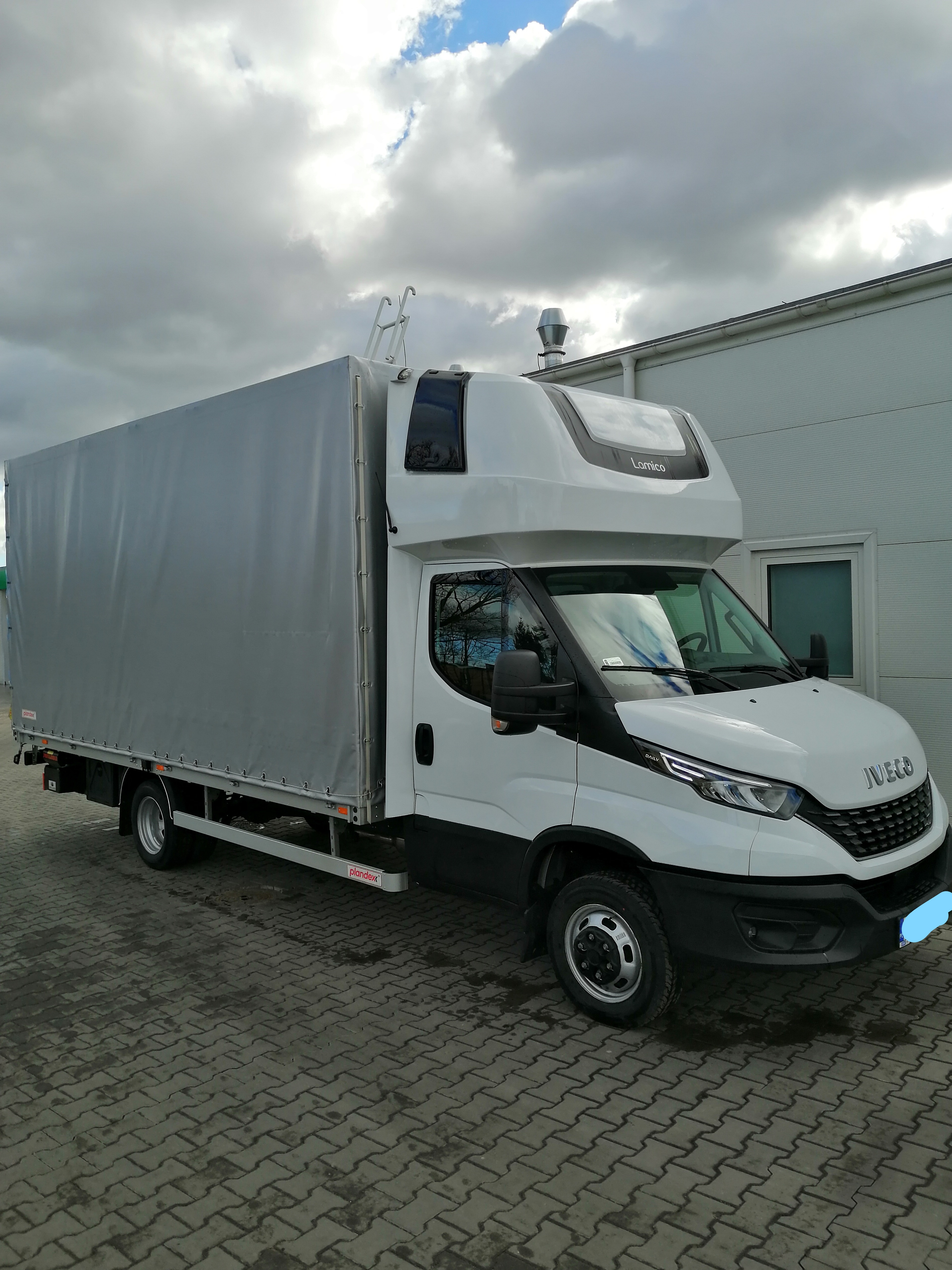 Biały samochód dostawczy Iveco Daily z plandeką na parkingu z kostki brukowej w pochmurny dzień.